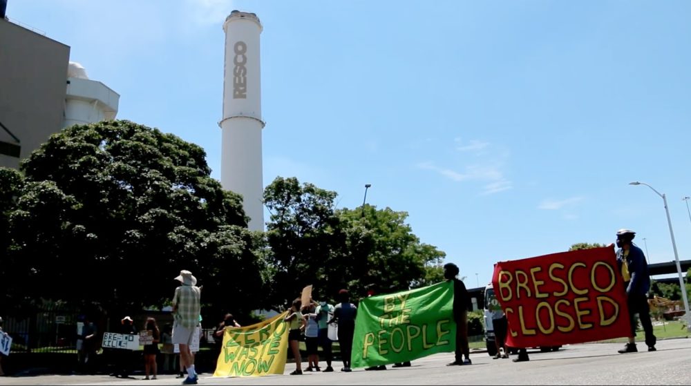 a group of people hold signs demonstrating against the Bresco trash incinerator, which is visible in the background