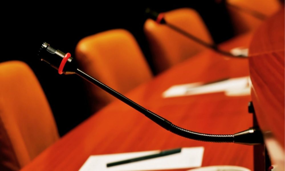 close-up of a table with empty chairs and speaker microphones, suggesting a meeting. notepads and pens are also on the table.