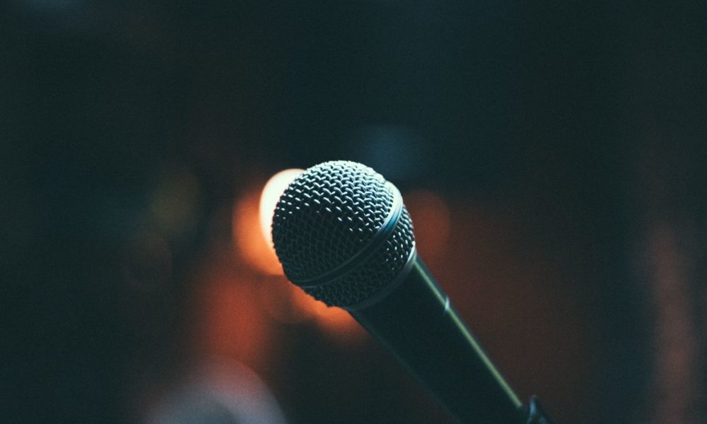 A microphone faces the viewer in the foreground; in the background, lights imply an expectant audience