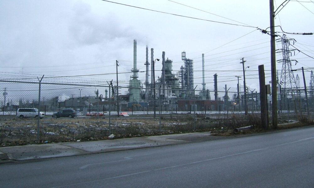 A grey sky blends with clouds of emissions from a petrochemical plant in Detroit, MI, seen through a chainlink fence