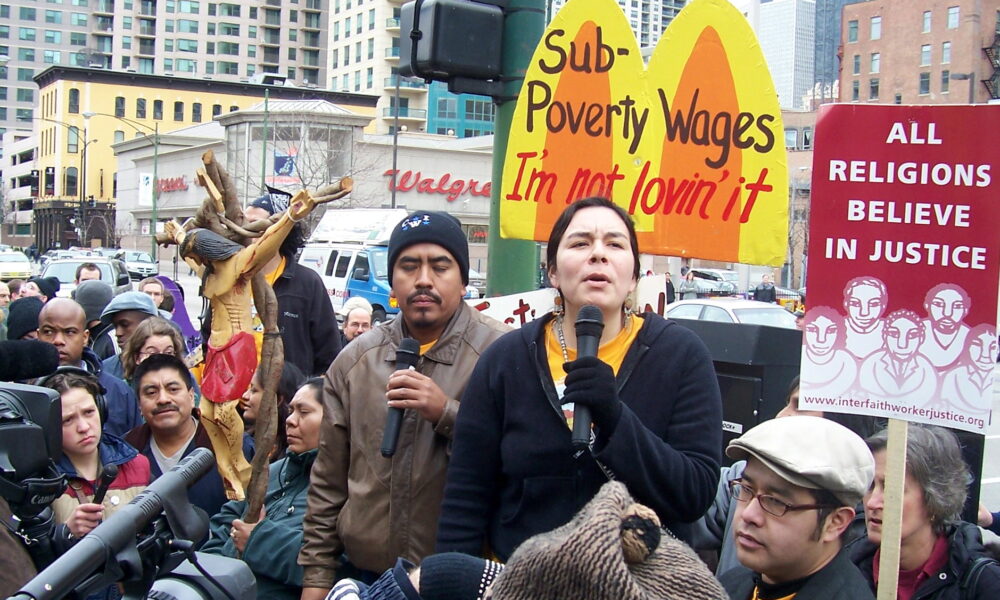 photo of a woman speaking to a crowd on a city street through a microphone, in front of a sign that says "sub-poverty wages: I'm not lovin' it"