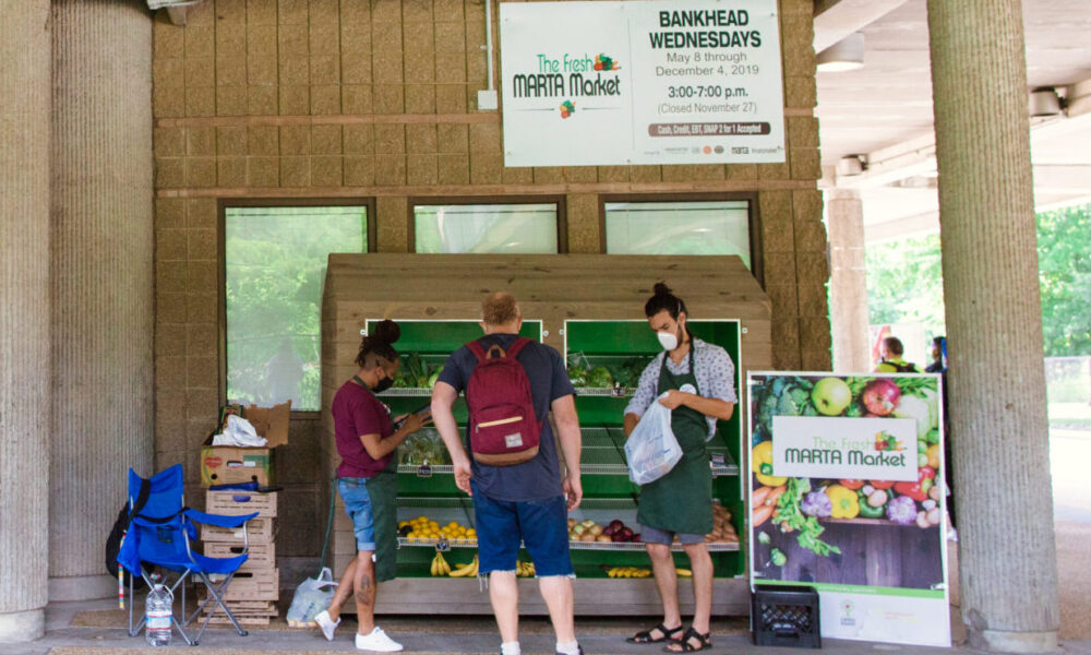 photo of a mobile farmers market set up at a commuter rail station; a man is reaching into his back pocket to pay for groceries that a masked employee is bagging while another masked employee rings up his purchase