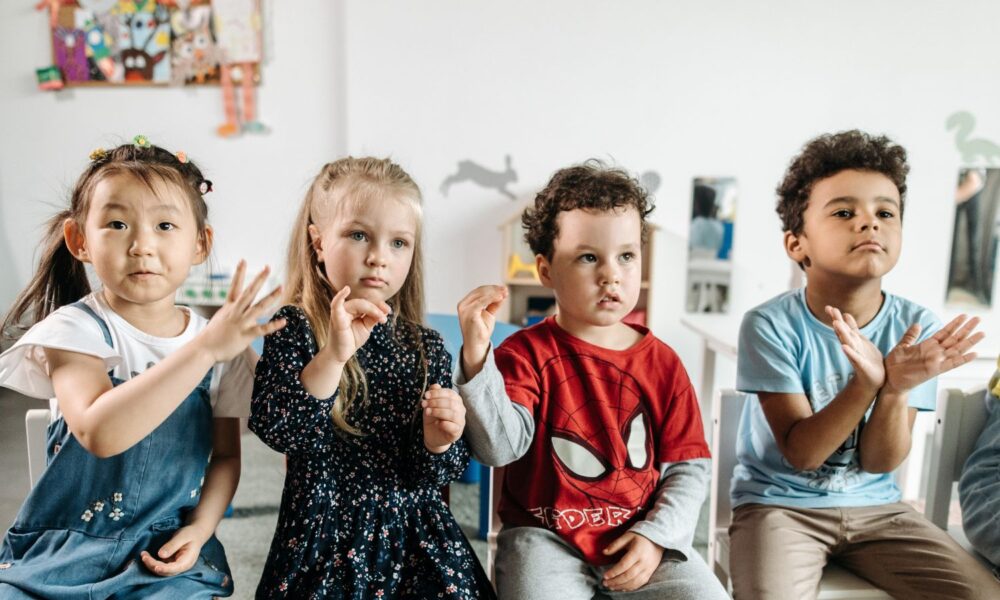 Four preschoolers of different races/ethnicities sit in a school holding their hands up as though they're reciting a song or lesson