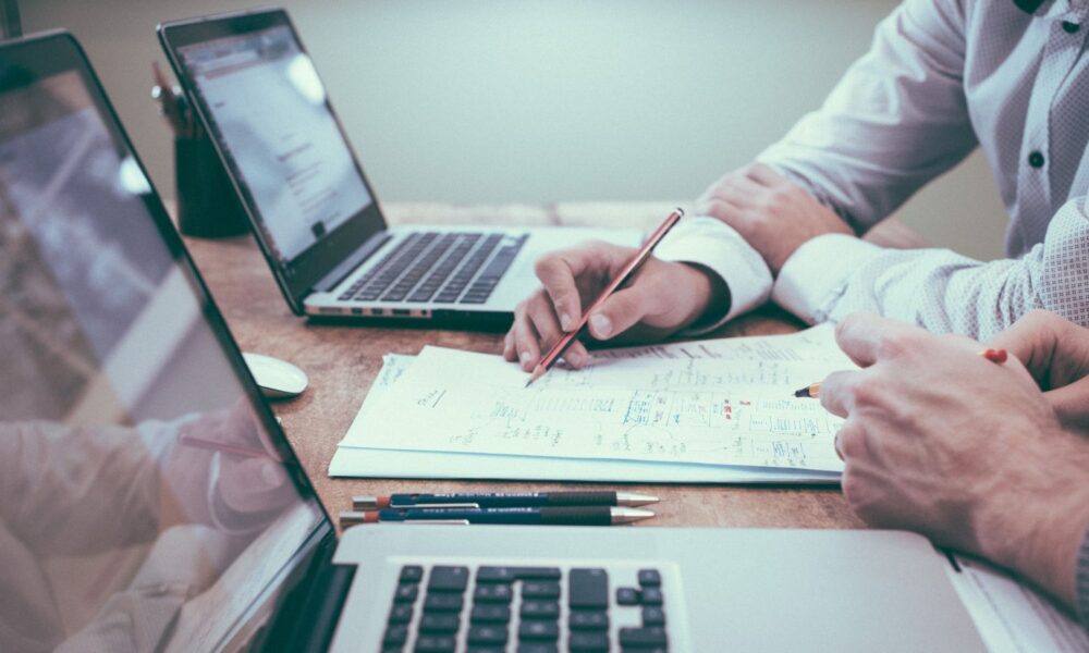 Two people sitting in front of two laptops. One holds a pencil over paper with calculations.