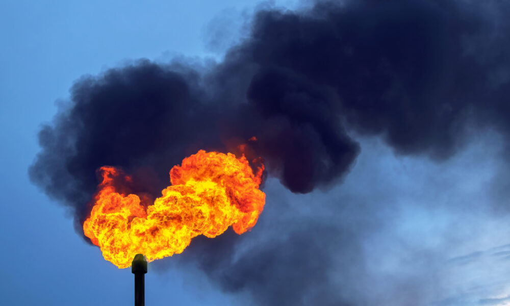 photo of a gas flare, with orange and red flames emanating from a stack and black smoke filling the blue sky