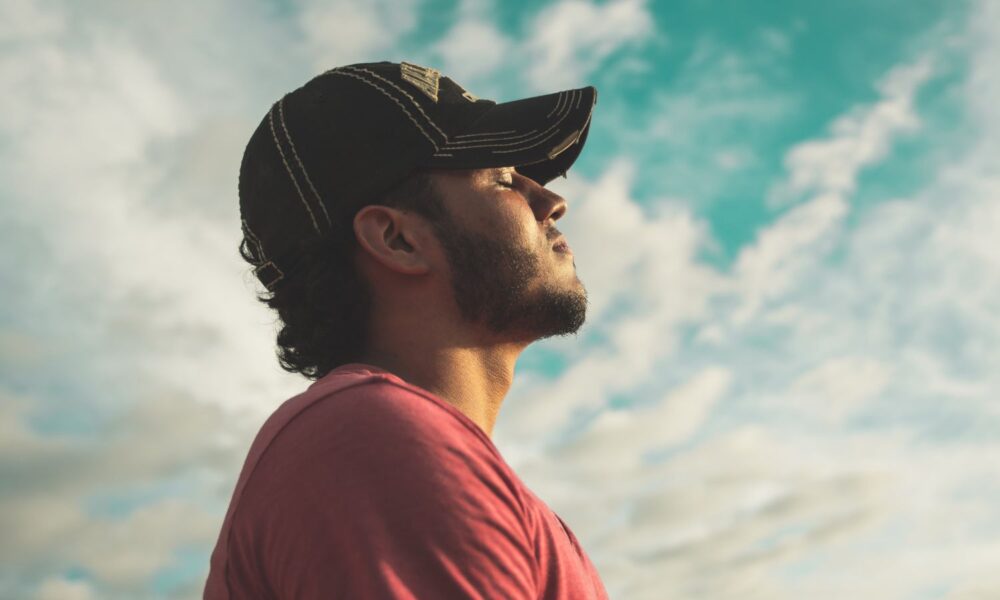 Man wearing black cap with eyes closed under cloudy sky.