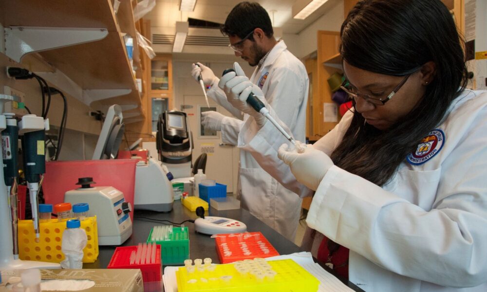 Two BIPOC FDA scientists wearing labcoats and protective gear prepare liquid containing nutrients that make it an ideal environment for growing bacteria. They are part of a group of researchers nicknamed “Team Tomato,” who try to understand and prevent Salmonella contamination early in the tomato production process.