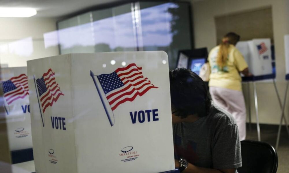 a woman sits at a booth to fill out a ballot. the booth says VOTE with a USA flag on it side.