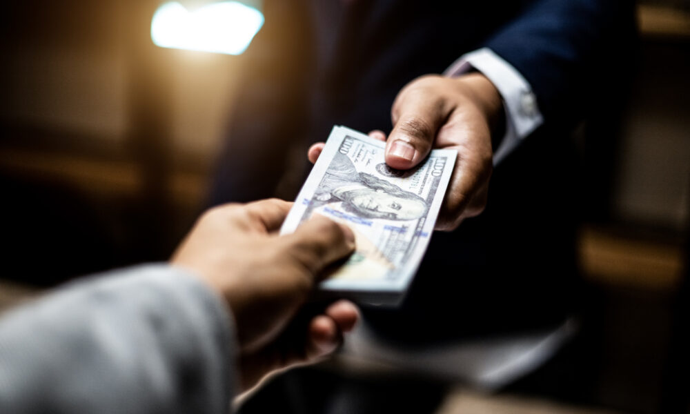 close-up photo of two men's hands exchanging a stack of $100 bills in a dimly lit room; one of the men is wearing a business suit