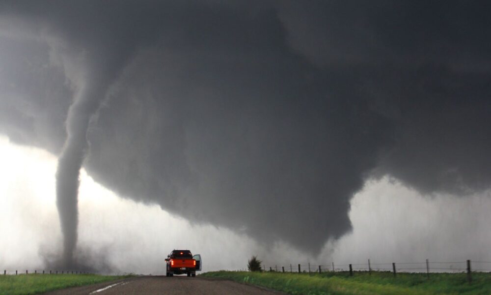 A red pickup truck idles on a road in rural Nebraska with its passenger side door open. Nearby, the massive black funnel cloud of a tornado touches down.