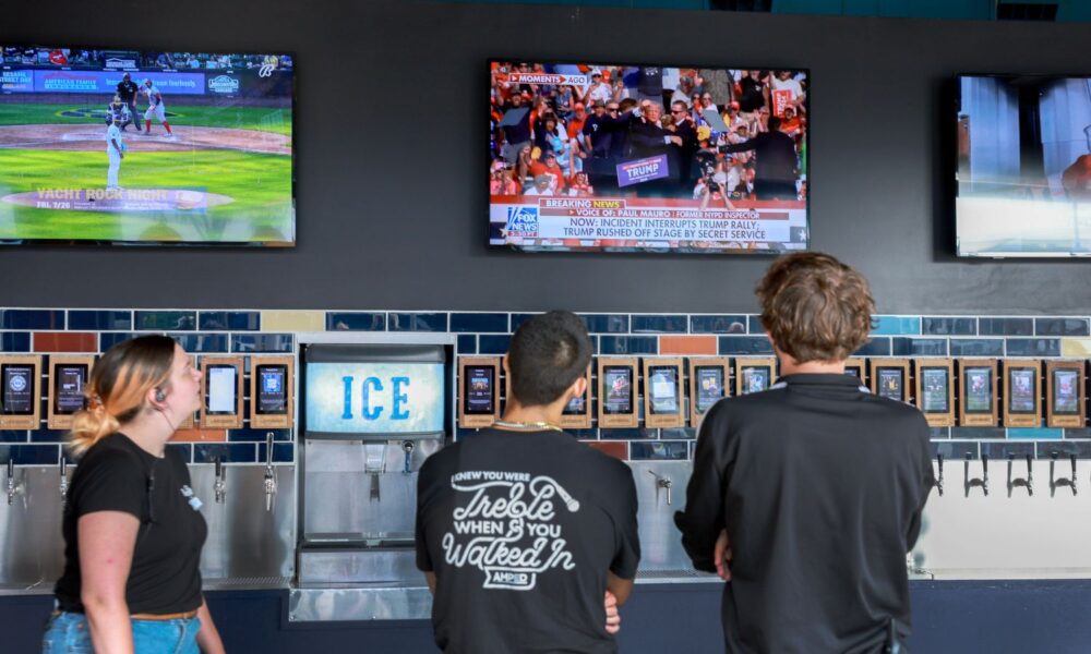 Three people watch televisions mounted above a bar: one screen shows a baseball game; the other shows footage of former president Trump holding his head after an assassination attempt at his rally in Pennsylvania in July 2024.