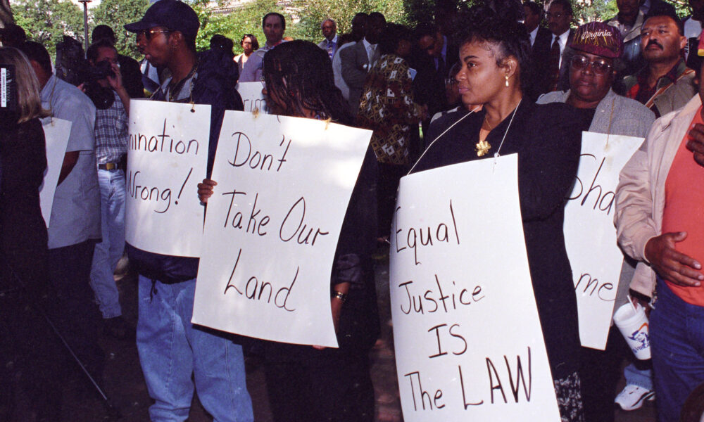 photo of male and female Black farmers at a protest, holding signs that read "Discrimination is wrong," "Don't take our land," and "Equal justice is the law"