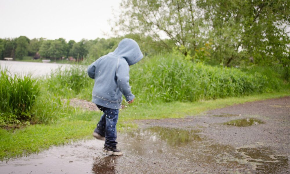 A kid wearing a gray hoodie, jeans, and sneakers splashes through a mud puddle outside on a gray day.