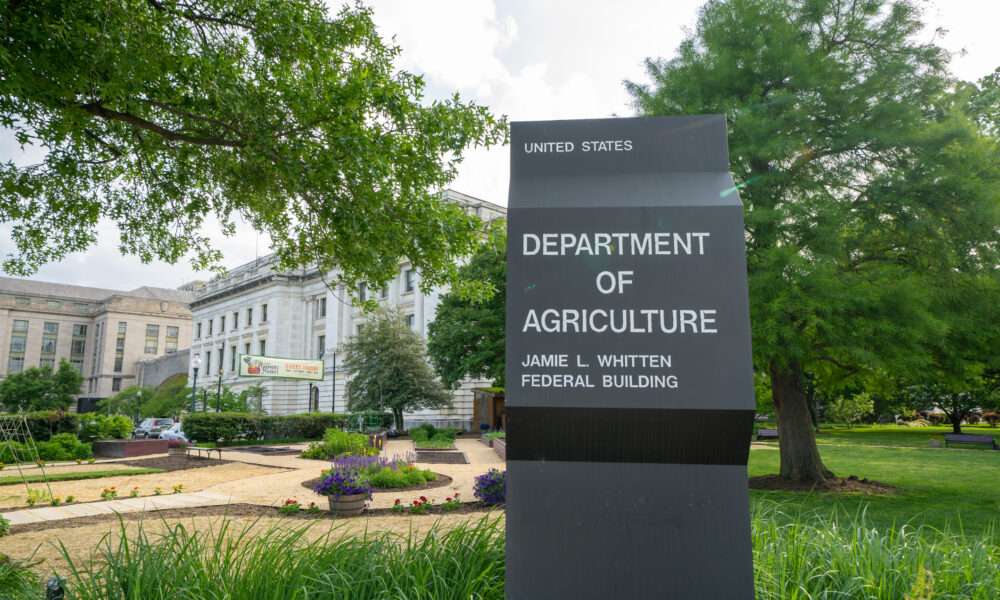 photo of sign for the US Department of Agriculture Whitten Building in Washington, DC; the building is in the background, among trees, a garden, and walkway