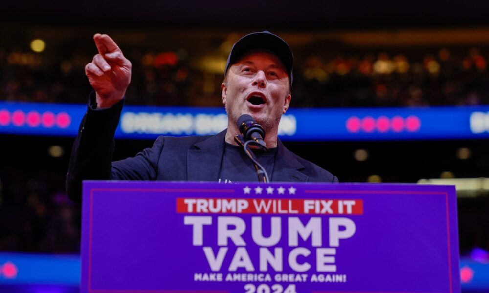 Elon Musk behind a podium with a Trump campaign sign speaks at a rally.