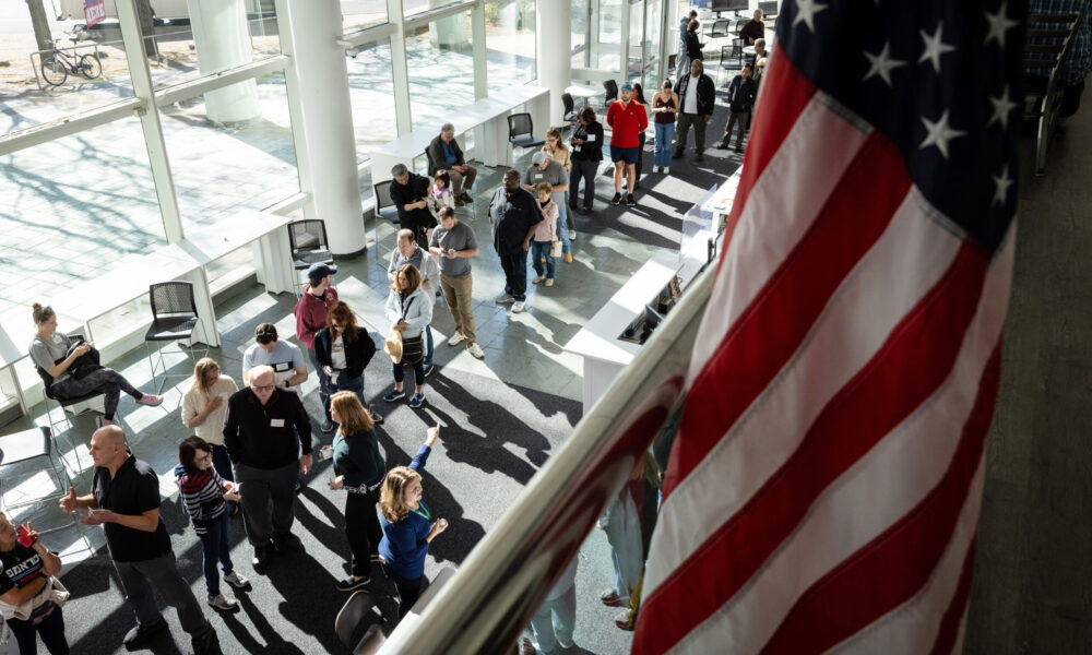 A shot from above of voters standing in a line, with an American flag in the foreground