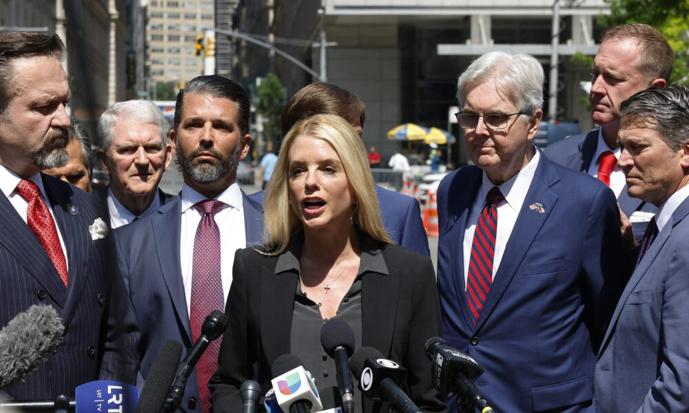 U.S. Attorney General nominee-designate Pam Bondi at the center of a group of people in suits, speaking at a microphone