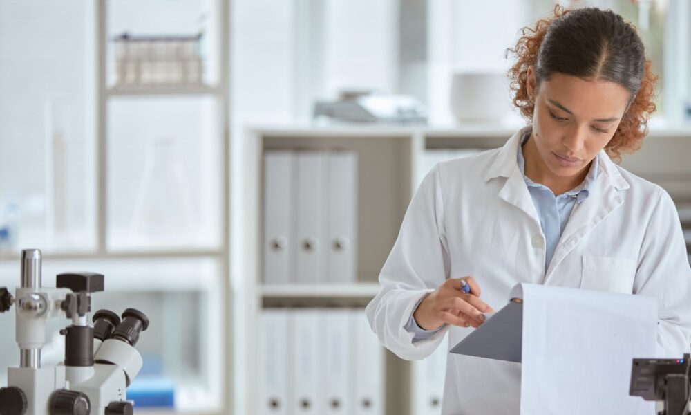 Female scientist reading notebook next to microscope in lab.