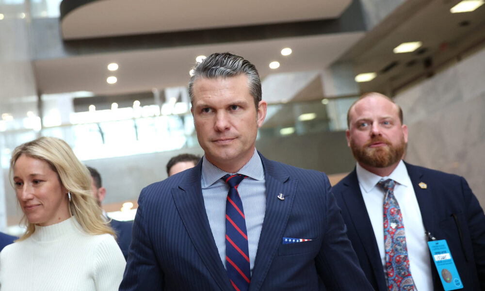 photo of Pete Hegseth walking down a hallway of a Senate office building, flanked by his wife to the left and an anonymous staffer to the right