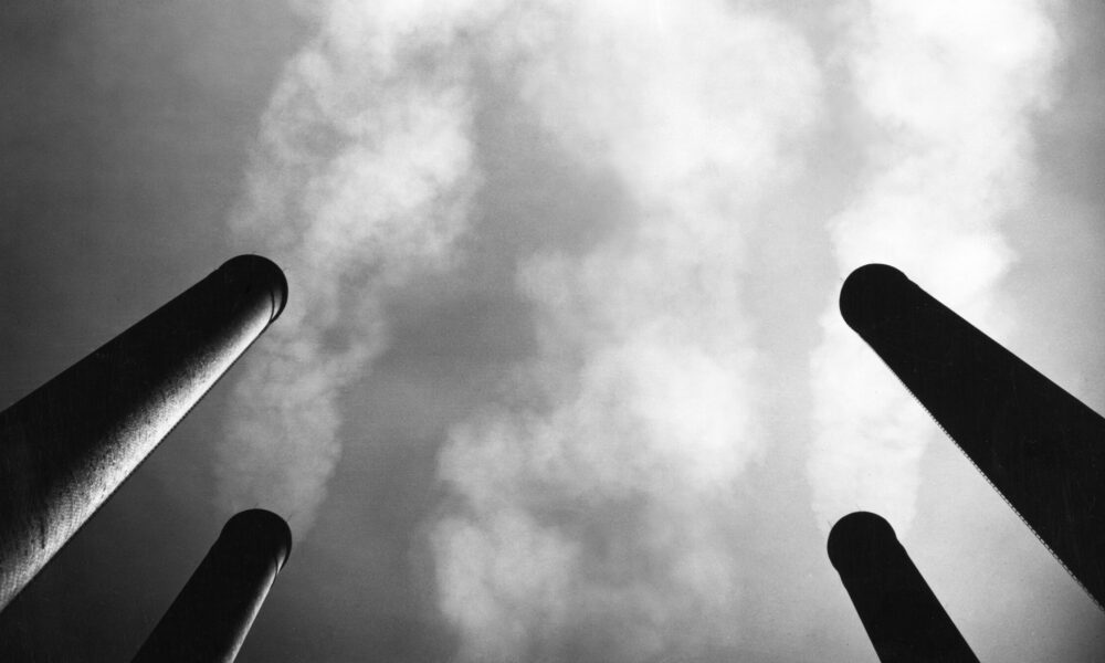 black and white photo of four smokestacks viewed from far below; smoke billows up into the sky from each one