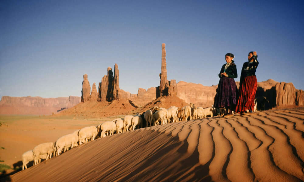 photo of two Navajo women wearing traditional clothing, standing on a sand dune and watching over a group of sheep; on the horizon, several tall rock pinnacles reach up into the blue sky