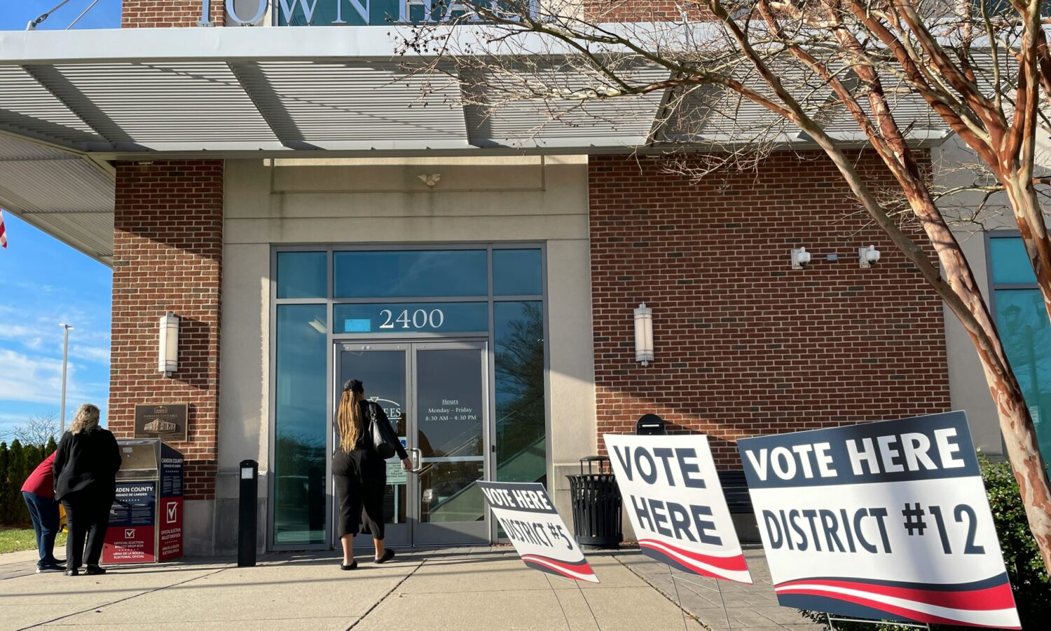 the exterior of a voting site, with two people entering and one using a ballot dropbox. three signs encouraging voting stand in the foreground.