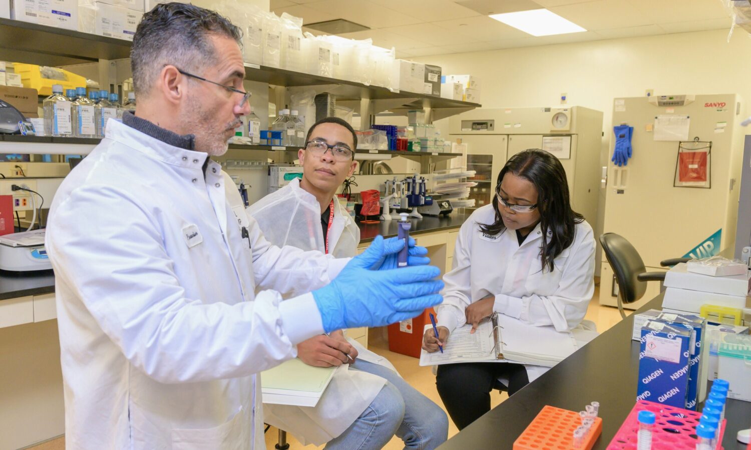 in a laboratory, two scientists in goggles and lab coats take notes as a third scientist speaks