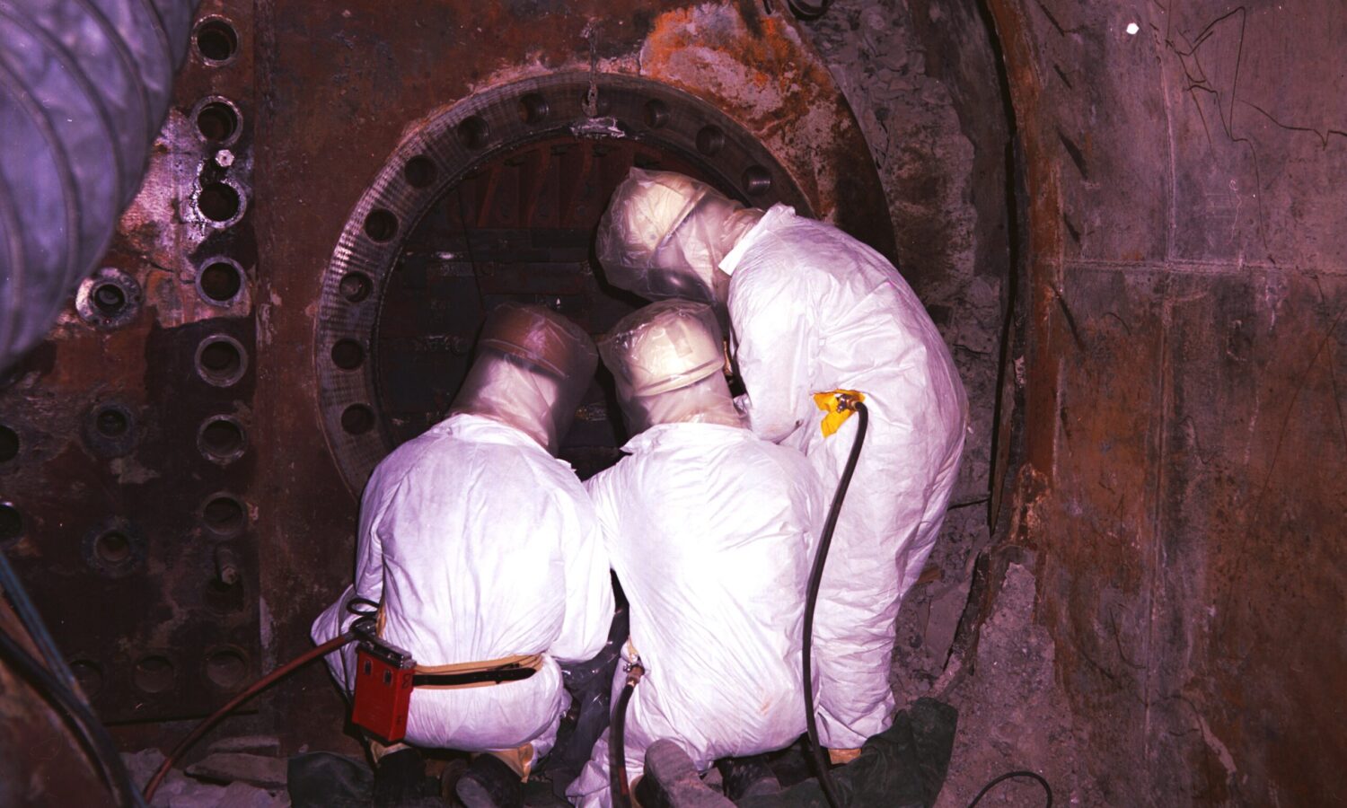 photo of three workers in white protective gear with their backs to the camera, collecting samples from an underground chamber following a nuclear test