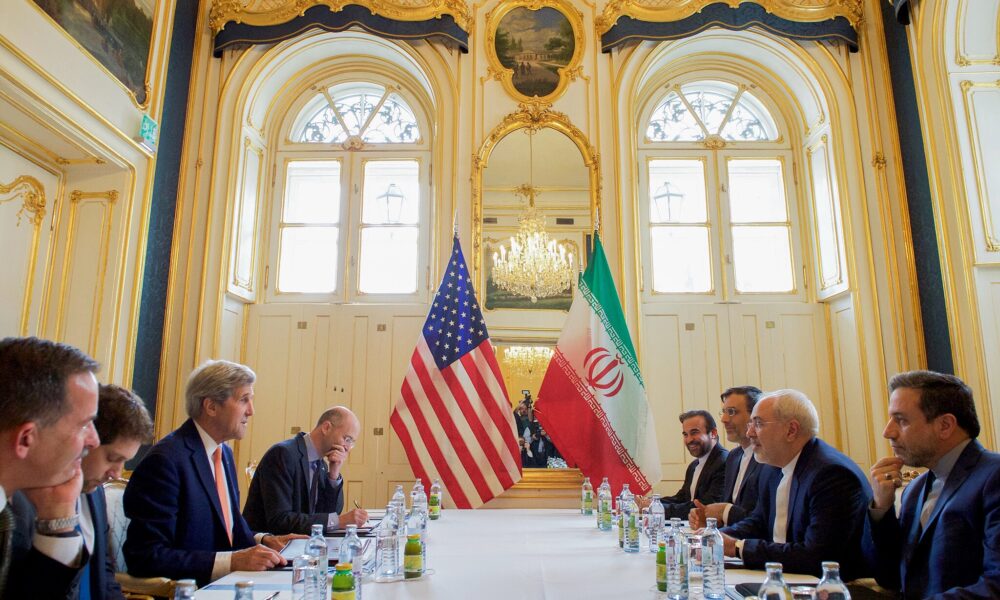 photo of US negotiators on the left side of a table and Iranian negotiators on the right side; the two countries' flags stand at the head of the table and light in shining into the high-ceiling room from two large windows above the flags