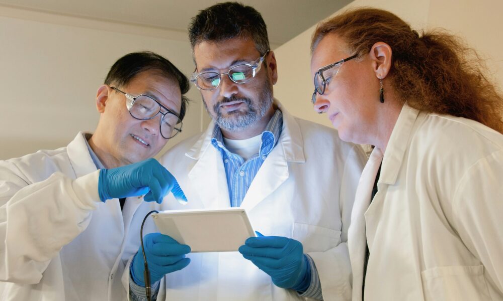three scientists in lab coats and goggles look at a tablet together