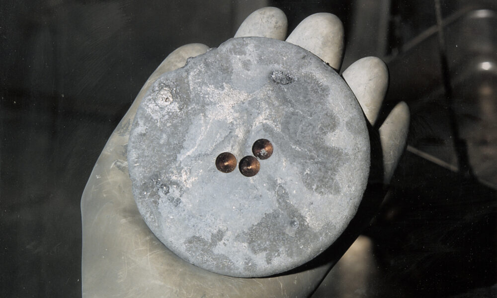 photo of a gloved hand holding a plutonium pit, which appears to be a gray metallic disc