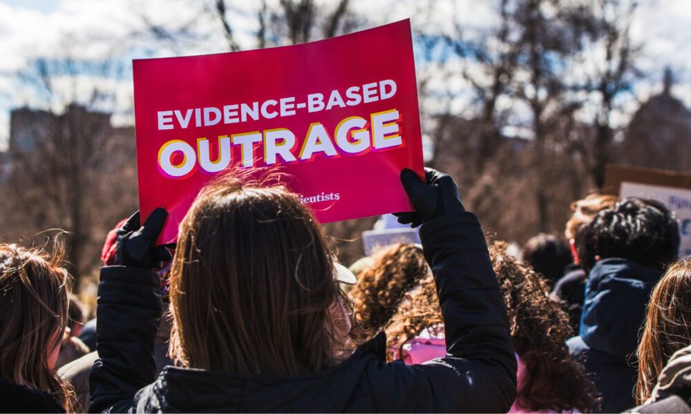 At a 2025 Stand up for Science rally in Boston, a person holds a sign reading "Evidence-Based Outrage"