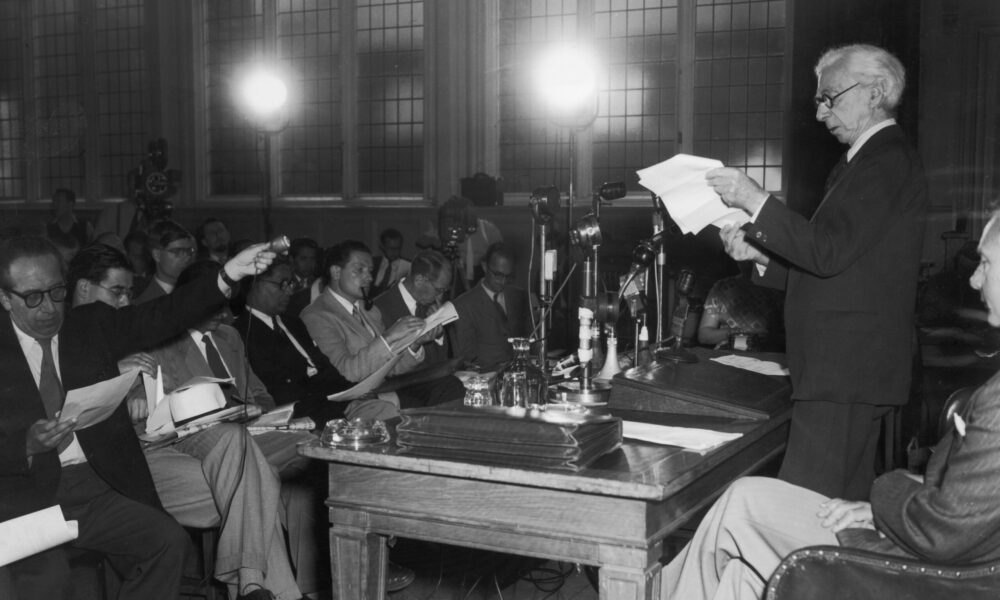 black-and-white photo of Bertrand Russell standing at a table and reading aloud from notes he is holding, with an audience of numerous reporters writing notes inside a darkened hall with bright lights in the background