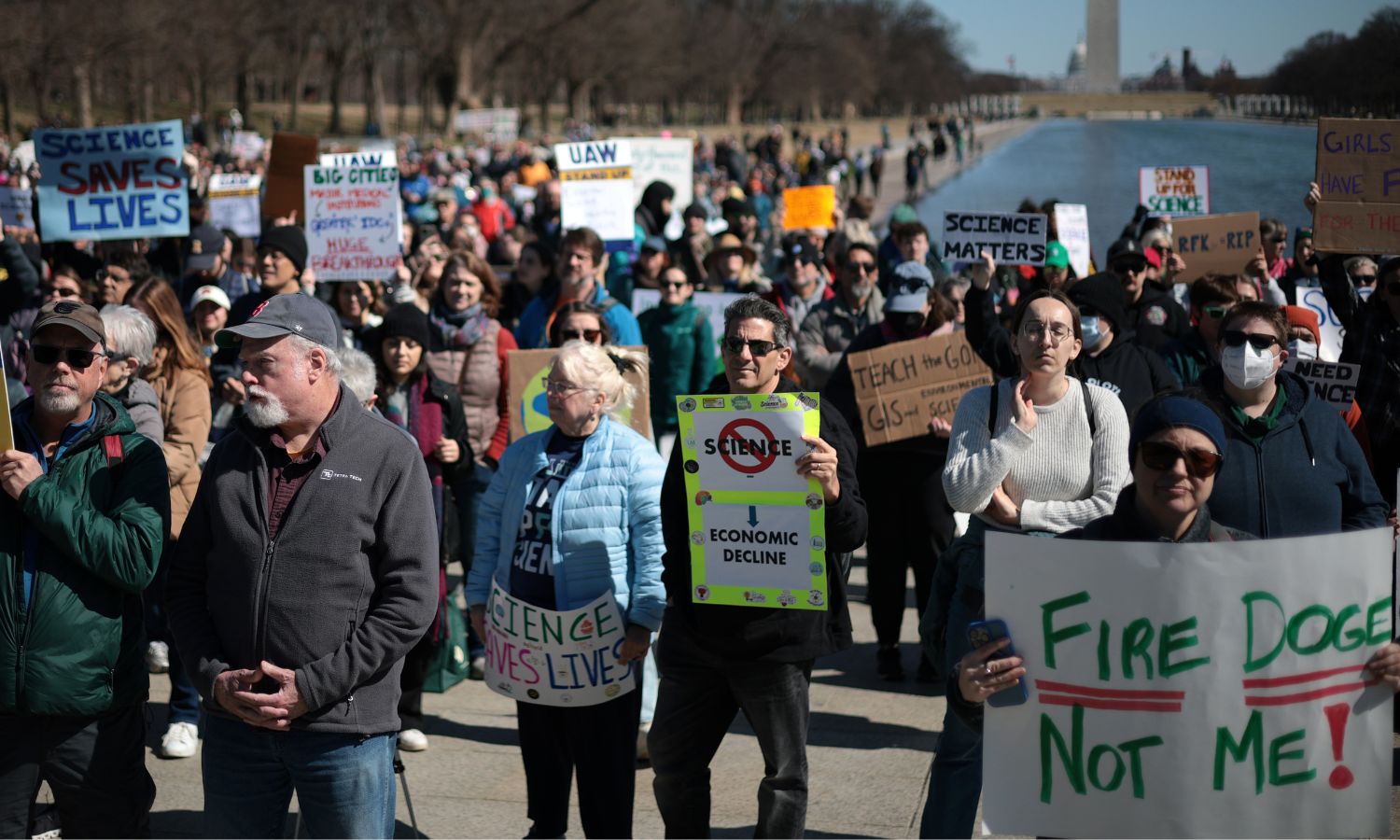 People holding signs rally in front of the Washington Monument in DC. One sign reads "Science Saves Lives"; another says "Fire DOGE, not me"
