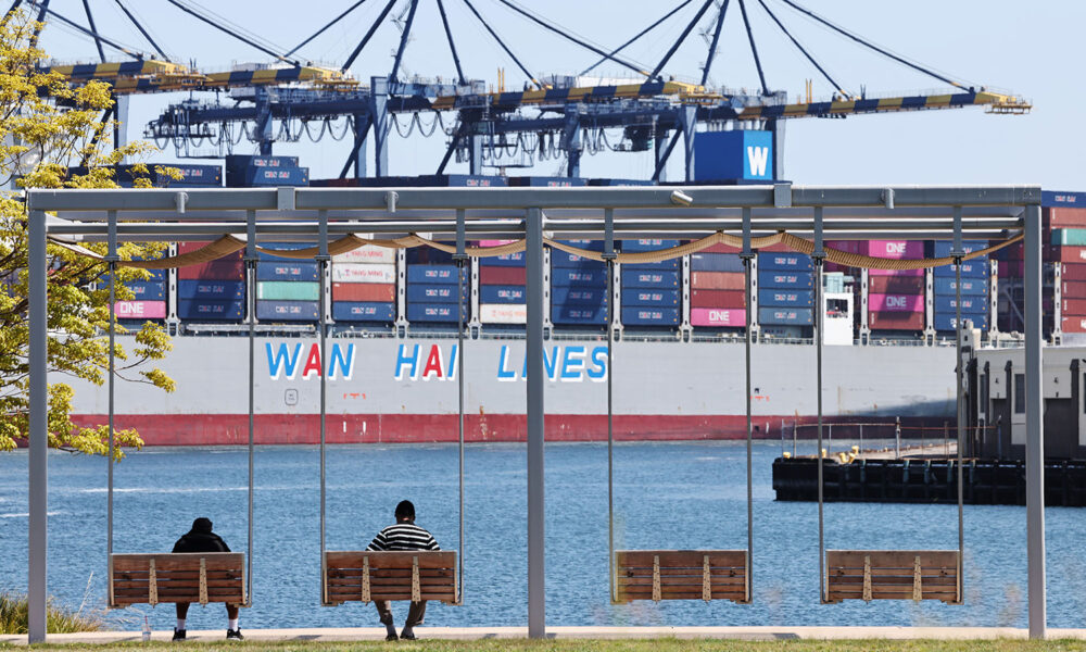 People sit in a park near shipping containers stacked on a container ship at the Port of Los Angeles