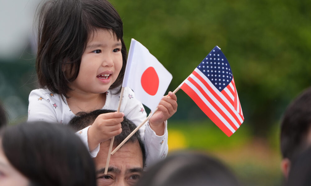 photo of a young smiling girl sitting on her father's shoulders and waving little US and Japanese flags