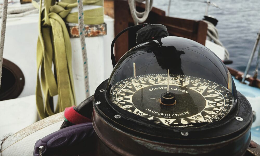 A traditional ships compass sits in the foreground of a photo taken in the cockpit of a wooden sailboat.