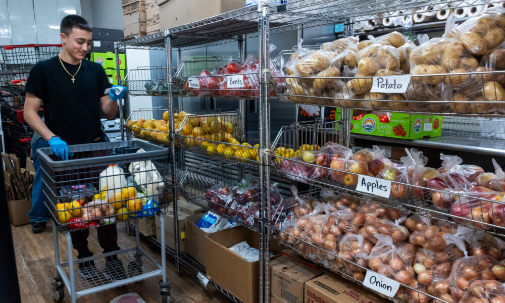 photo of a young man reading a shopping list while pushing a cart through the aisle of a food pantry. The shelves in front of him are labeled "Potato," "Apples," and "Onion."