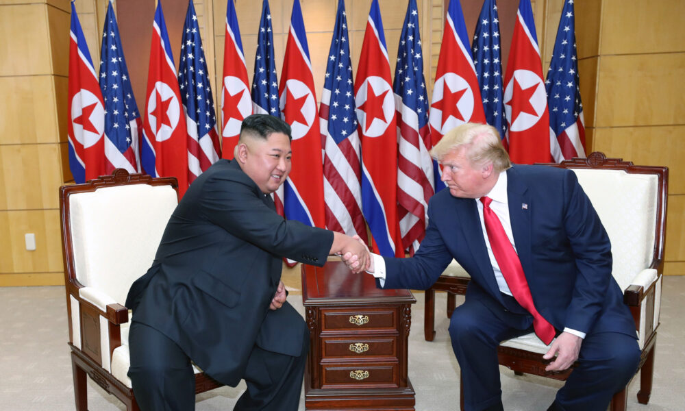 photo of Kim Jong Un and Donald Trump seated and leaning in toward each other to shake hands. A row of alternating US and North Korean flags is in the background.