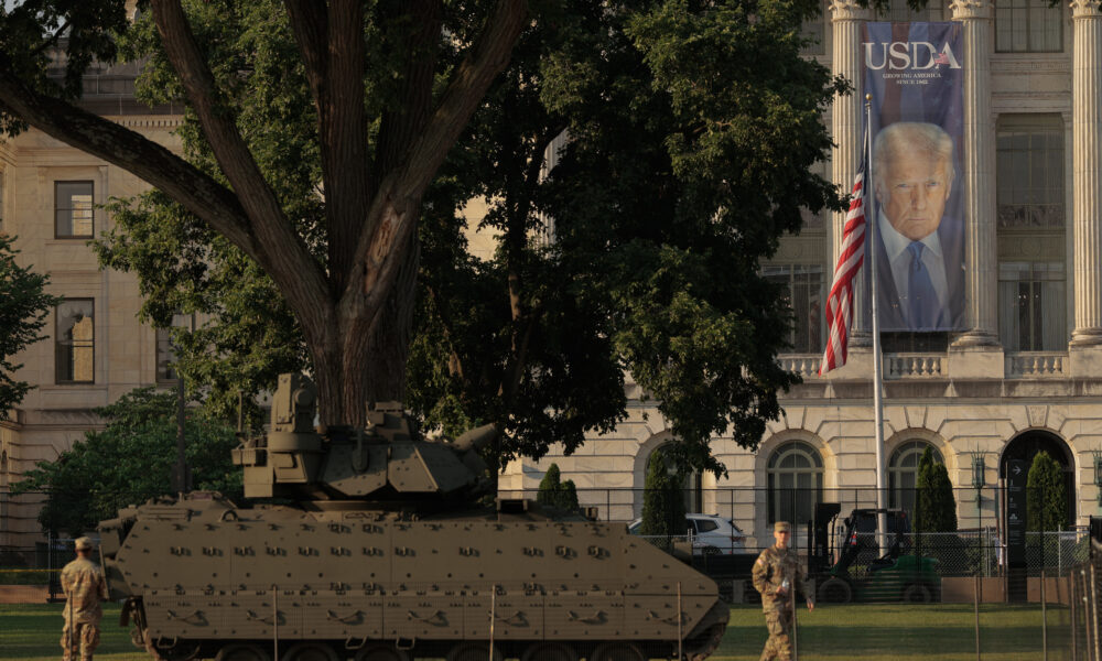 photo of two US Army soldiers and a Bradley fighting vehicle parked in front of USDA headquarters, on which hangs a long banner featuring an image of President Donald Trump