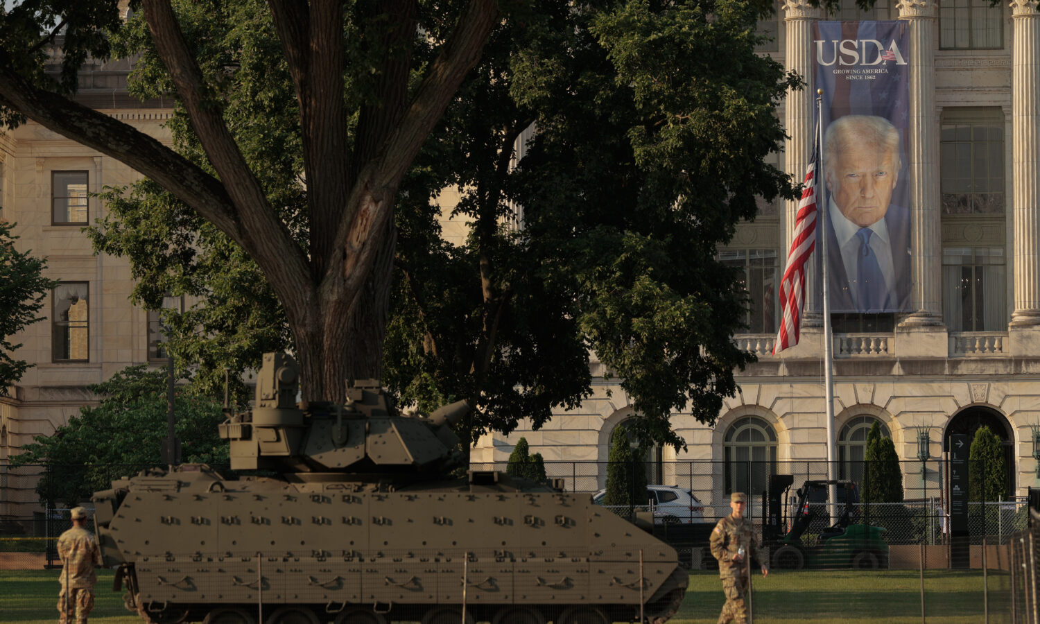 photo of two US Army soldiers and a Bradley fighting vehicle parked in front of USDA headquarters, on which hangs a long banner featuring an image of President Donald Trump