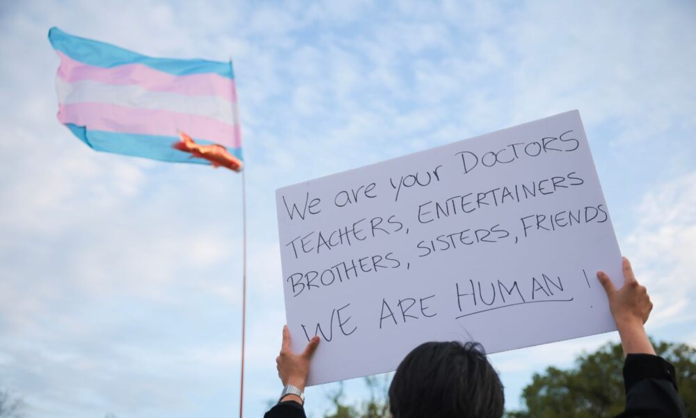 A person holds a sign reading "we are your doctors, teachers, entertainers, brothers, sisters, friends. WE ARE HUMAN!" as they attend a Washington, DC, rally supporting Trans Day of Visibility in 2025.