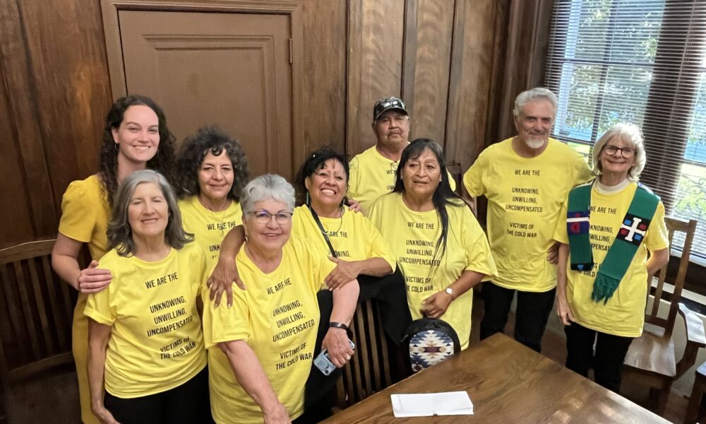 group photo of advocates smiling after their success in getting RECA passed; they all wear matching yellow T-shirts that read, "We are the unknowing, unwilling, uncompensated victims of the Cold War"