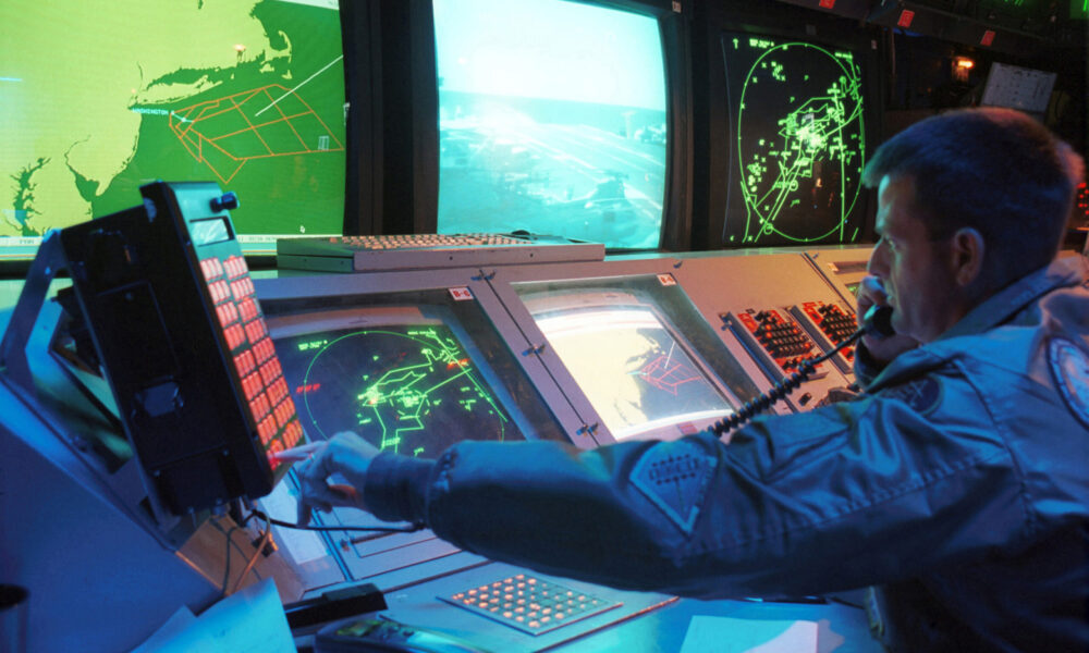 photo of a US naval officer monitoring the air space around New York City; he has a phone in one hand and is pressing a button on a lit panel with the other hand. There are several displays on the desk in front of him and larger screens on the wall beyond the desk