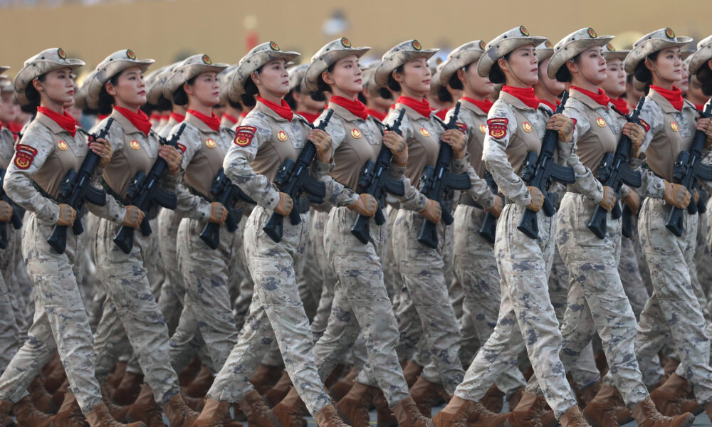 photo of an all-female unit of Chinese troops marching during a military parade; they wear matching gray camouflage uniforms with red scarves and all carry automatic weapons