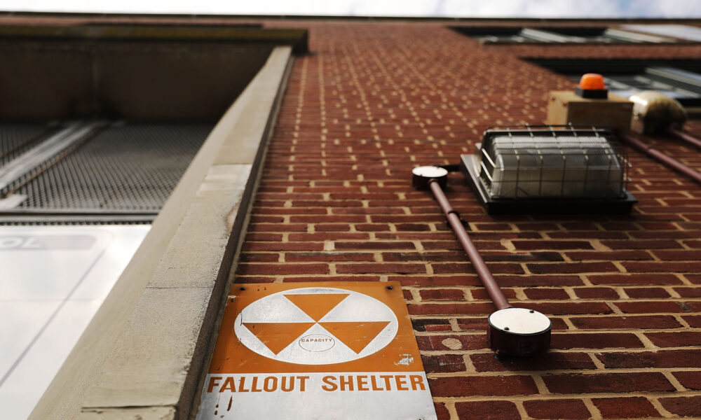 photo looking up at a brick wall, at the bottom of which is an old sign with a radiation symbol and the words "fallout shelter"