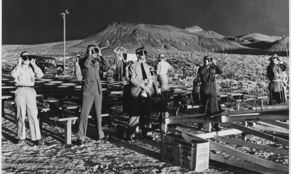 black and white photo of people observing a nuclear test explosion in the Nevada desert in 1955; they all hold goggles to their faces to protect themselves from the flash of the blast