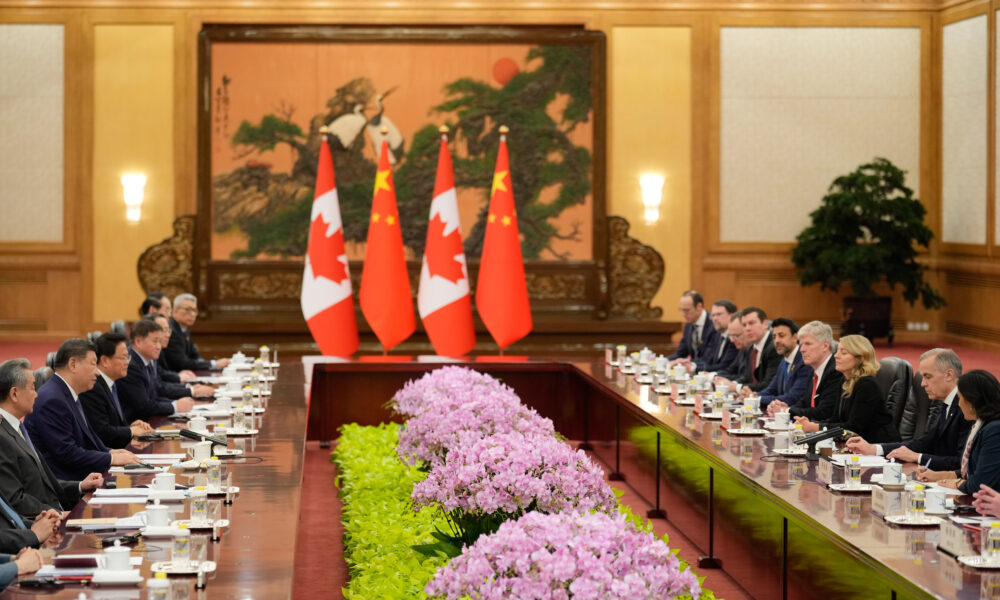 photo of the Chinese-Canadian summit meeting in Beijing, January 2026; the Chinese delegation sits at a table on the left; the Canadian delegation sits at a table on the right; there are flowers set between the tables and both countries' flags in the background between tables