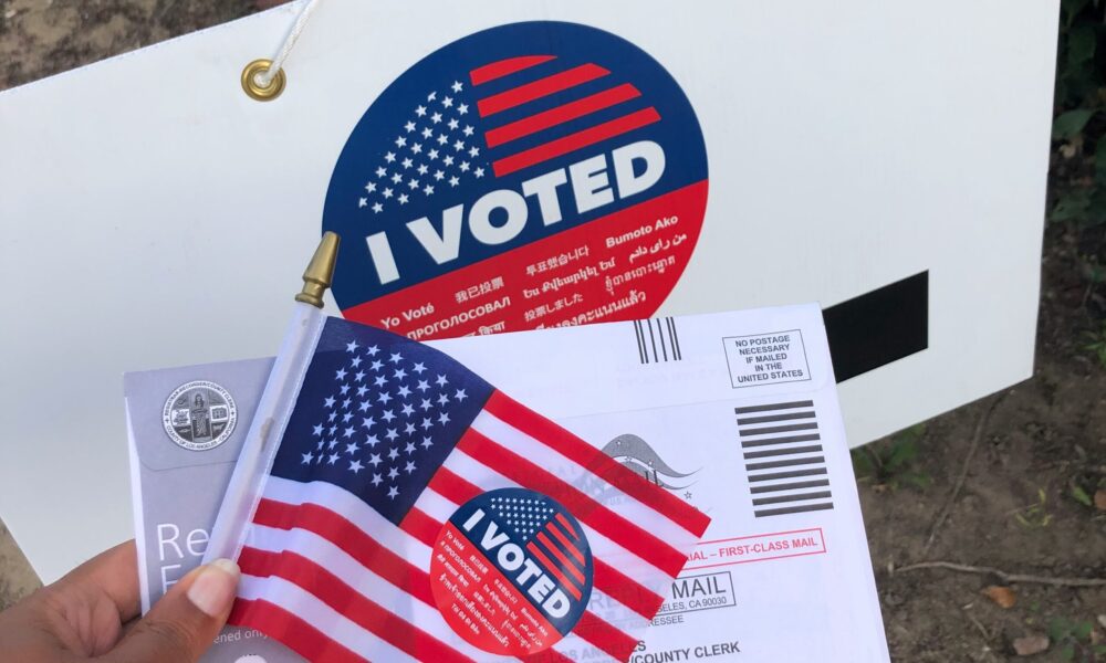 a hand holding a mail ballot and a miniature American flag in front of a yard sign that says "I Voted"