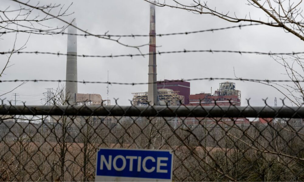 A blue sign reading NOTICE is attached to a fence in front of a coal-fired power plant spewing emissions into a gray sky over Harrodsburg, Kentucky. Under President Trump, EPA administrator Lee Zeldin revoked a finding that carbon dioxide emissions are hazardous to human health.